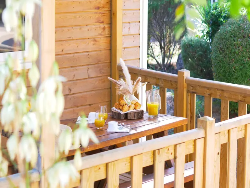 Une table en bois avec des viennoiseries et des jus d'orange est mise en scène sur une terrasse en bois.