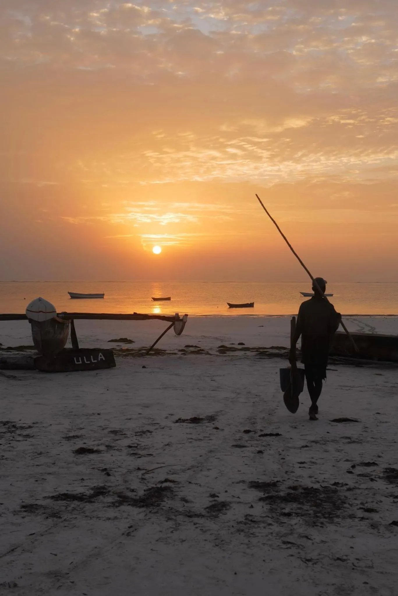 Un homme marche sur une plage au coucher du soleil, avec des bateaux à l'horizon.