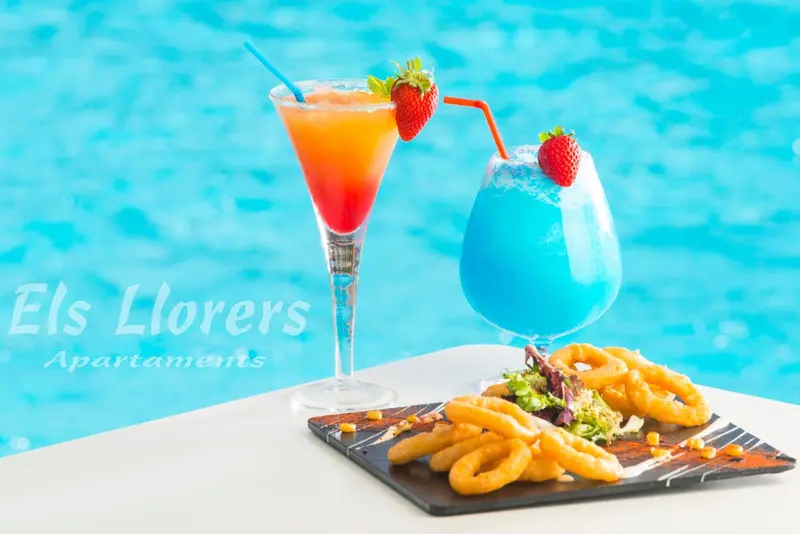 Deux cocktails colorés et des amuse-gueules sont présentés sur une table blanche au bord d'une piscine.