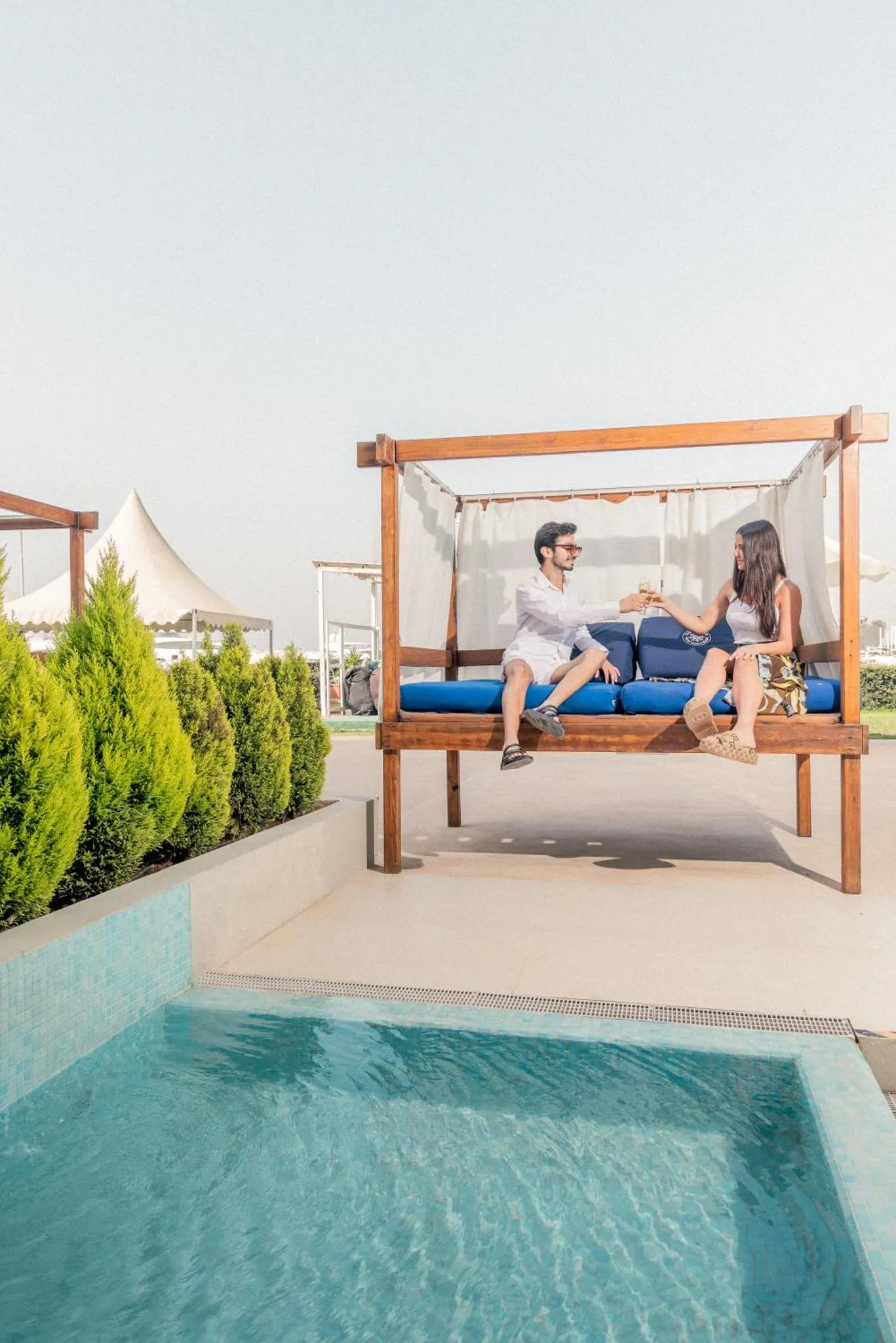 Un couple se détend sur une banquette en bois à l'ombre d'un auvent près d'une piscine.