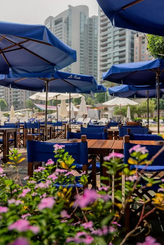 Tables et chaises bleues sous des parasols, avec une végétation fleurie au premier plan.