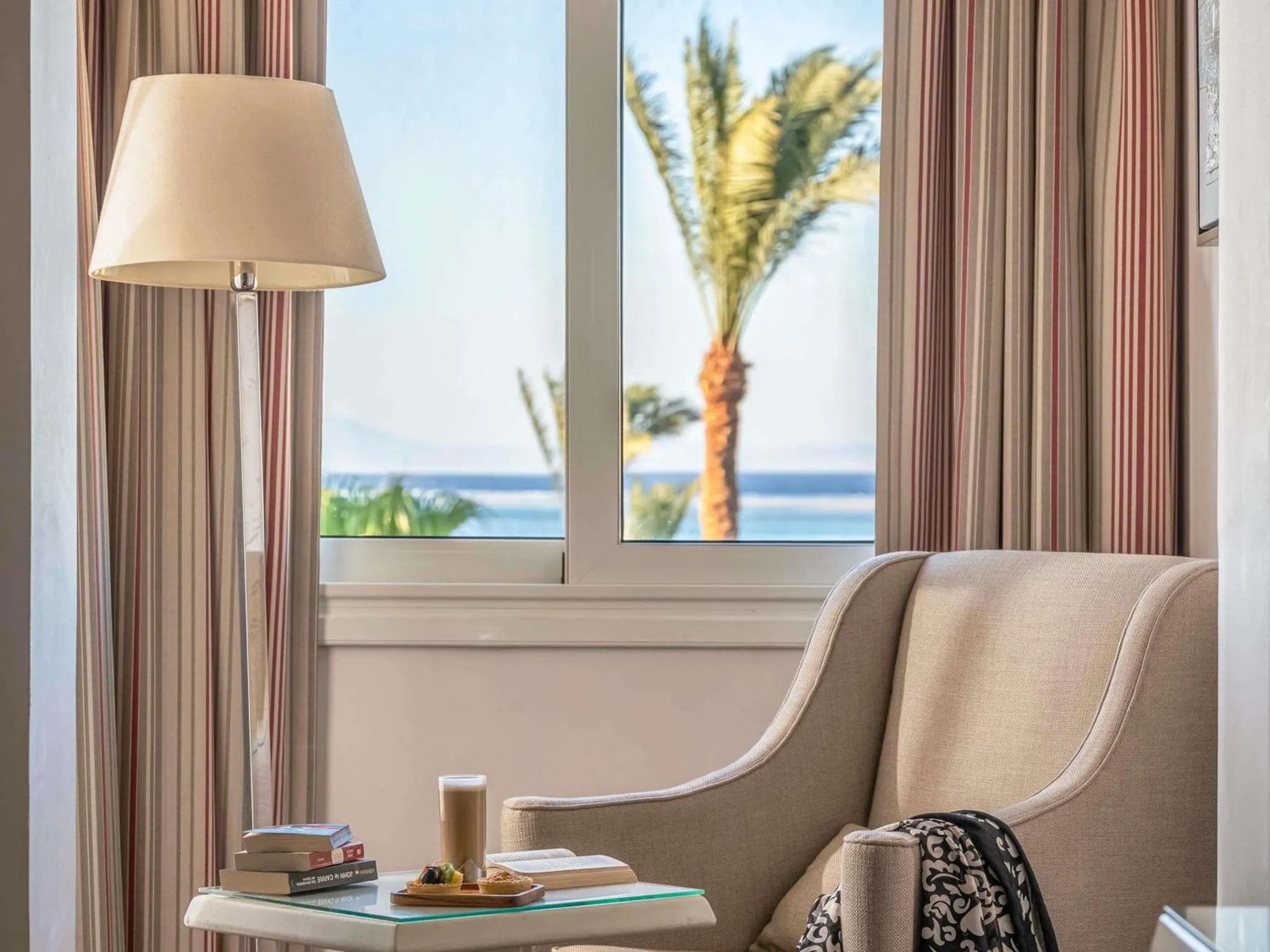Une chaise et une table basse avec une boisson et des livres sont placés devant une grande fenêtre donnant sur la mer.
