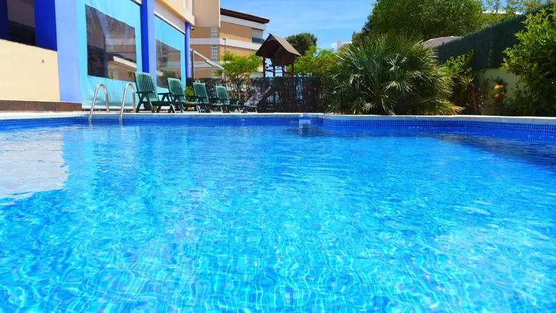 Une piscine rectangulaire bleu clair avec des transats verts sur une terrasse ensoleillée.