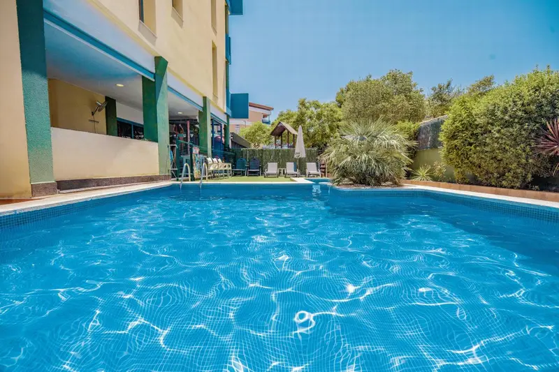 Une piscine rectangulaire turquoise est au premier plan, entourée d'une terrasse carrelée.