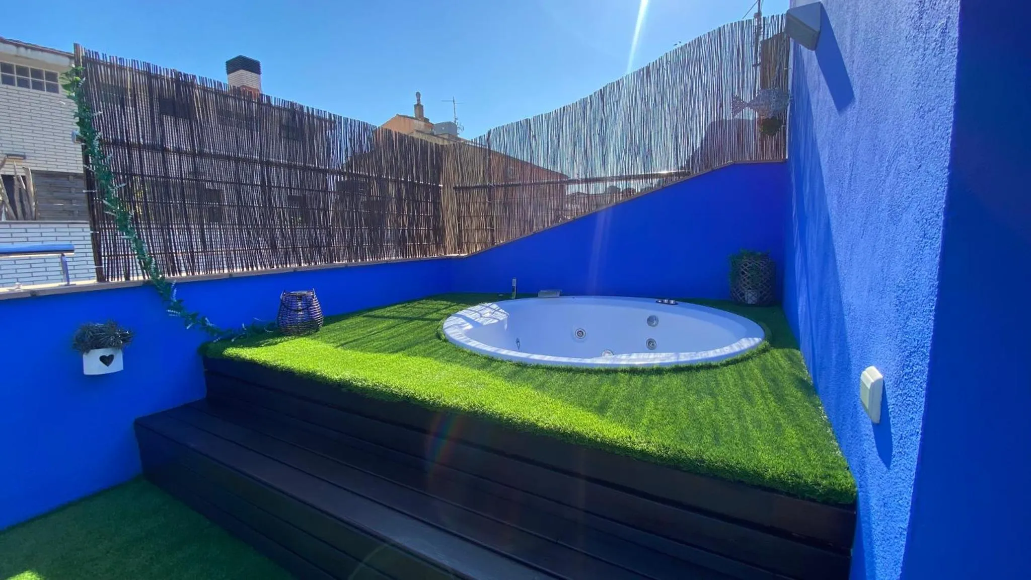 Un jacuzzi rond est installé sur une terrasse avec gazon artificiel et un mur bleu.