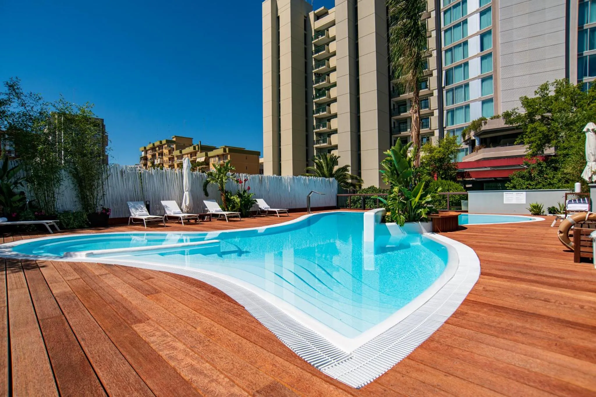 Piscine extérieure avec terrasse en bois et bâtiments en arrière-plan.