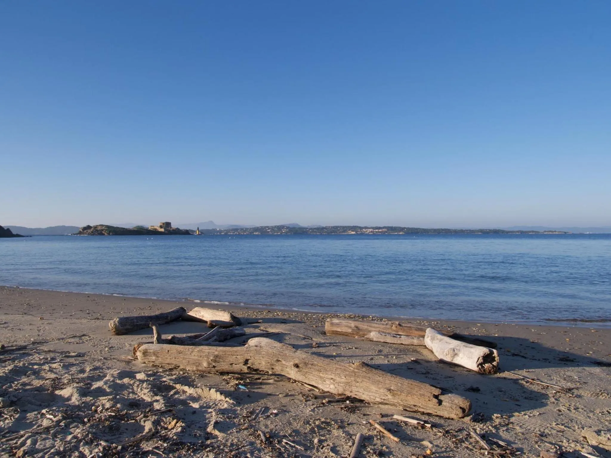 Plage de sable avec des troncs d'arbres échoués et une vue sur la mer et les îles au loin.