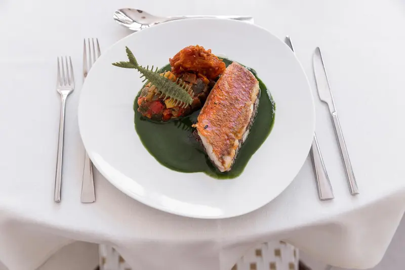 Assiette de poisson grillé avec accompagnements et sauce verte, couverts disposés sur une nappe blanche.