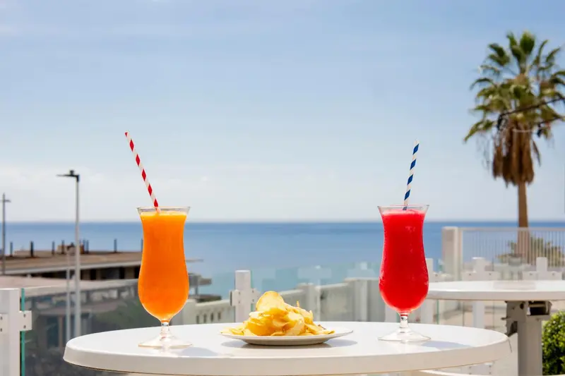 Deux cocktails colorés et des chips sont servis sur une table blanche en terrasse avec vue sur la mer.