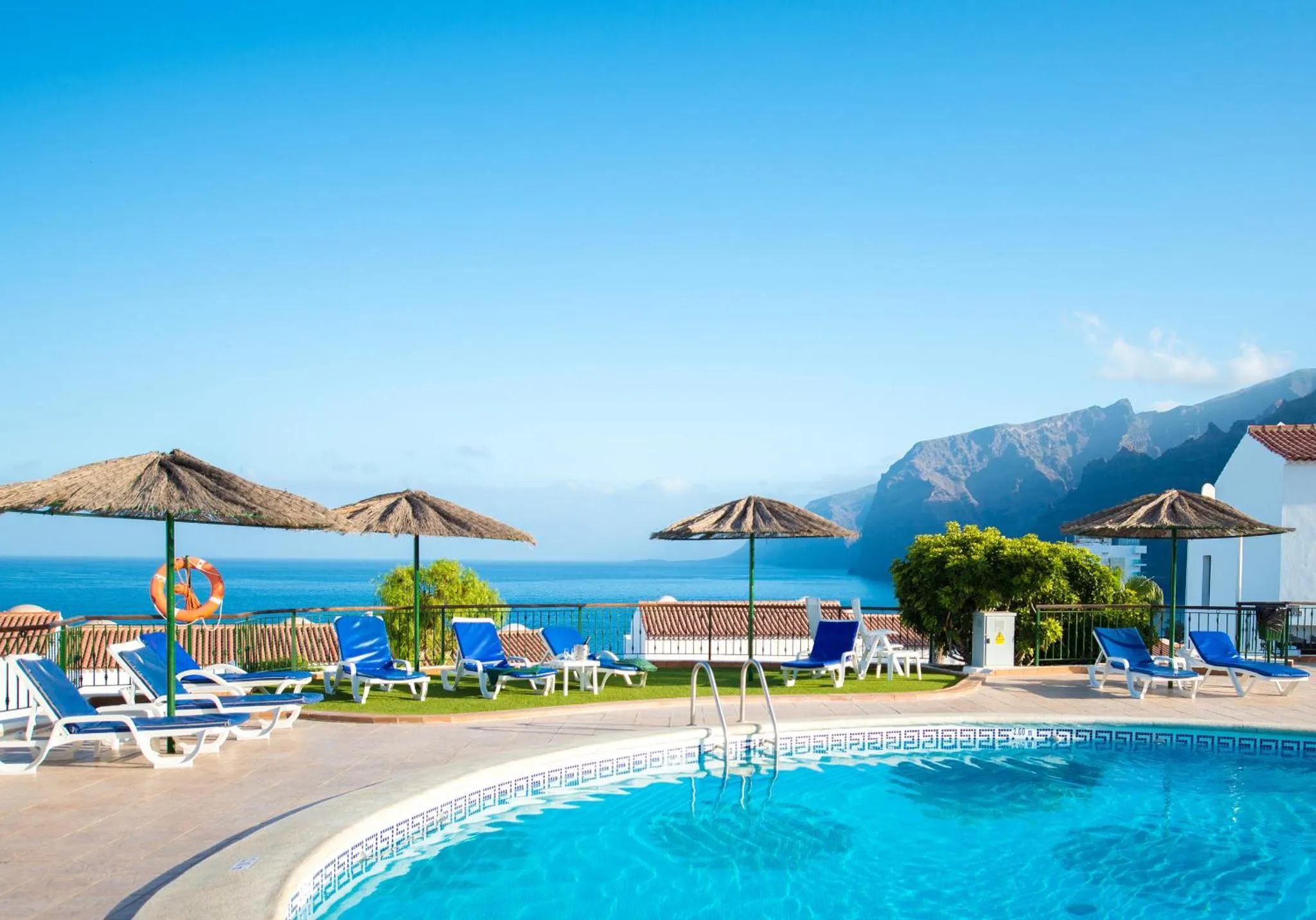 Transats bleus et parasols sur le bord d'une piscine turquoise avec vue sur la mer et les montagnes.