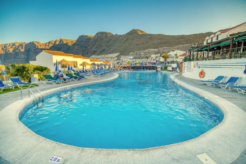 Une piscine rectangulaire bordée de transats et de parasols, avec des montagnes en arrière-plan.
