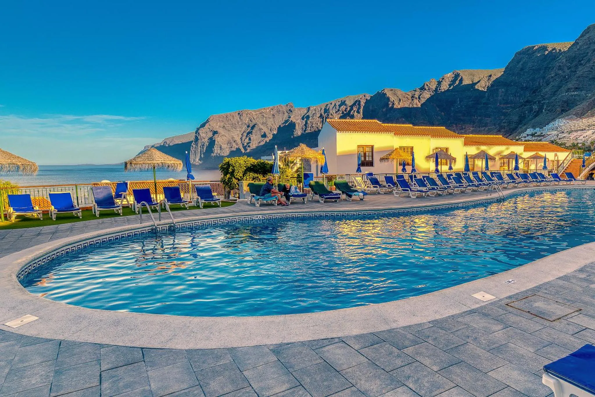 Piscine bleue bordée d'une terrasse avec transats et parasols, face à la mer et aux montagnes.