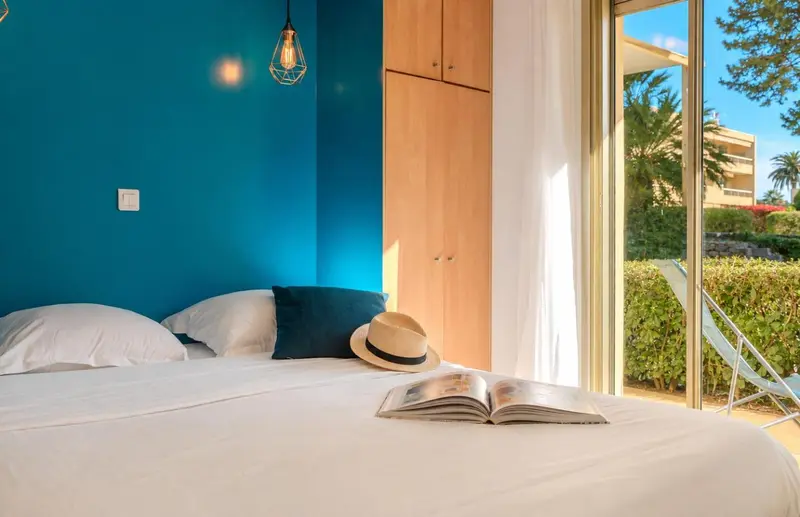 Un lit blanc avec un livre ouvert et un chapeau de paille, dans une chambre lumineuse.