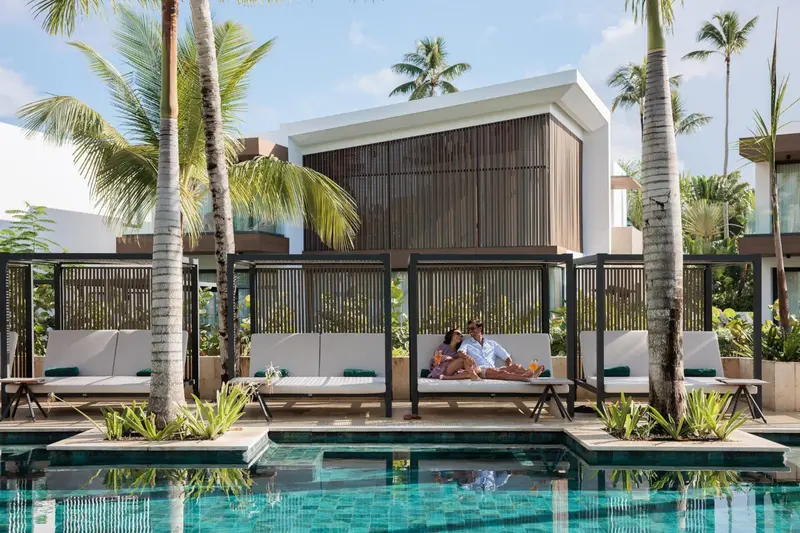 Des transats blancs sont disposés au bord d'une piscine turquoise, devant des bâtiments modernes.