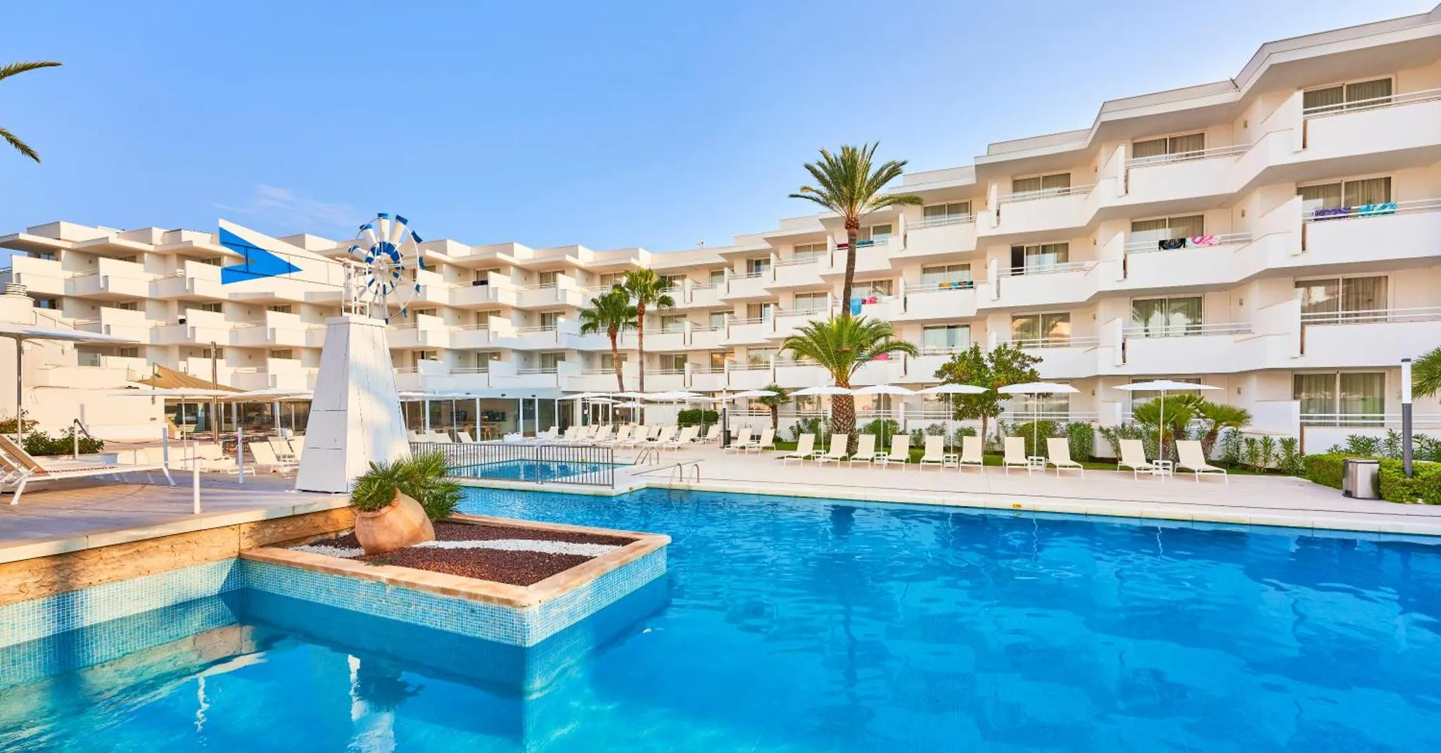 Vue d'une piscine devant un bâtiment blanc avec des transats et des palmiers.