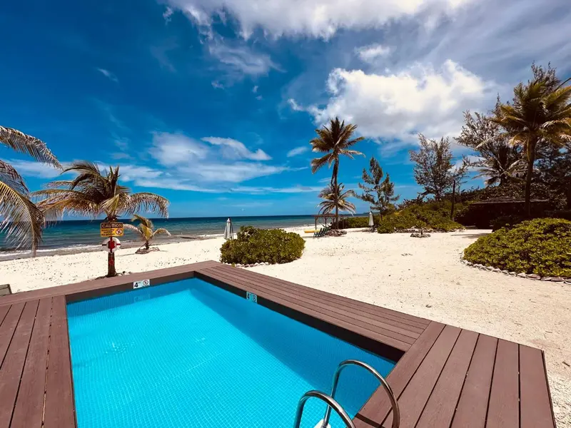 Petite piscine turquoise bordée de bois, sur une plage de sable blanc avec palmiers et mer bleue.