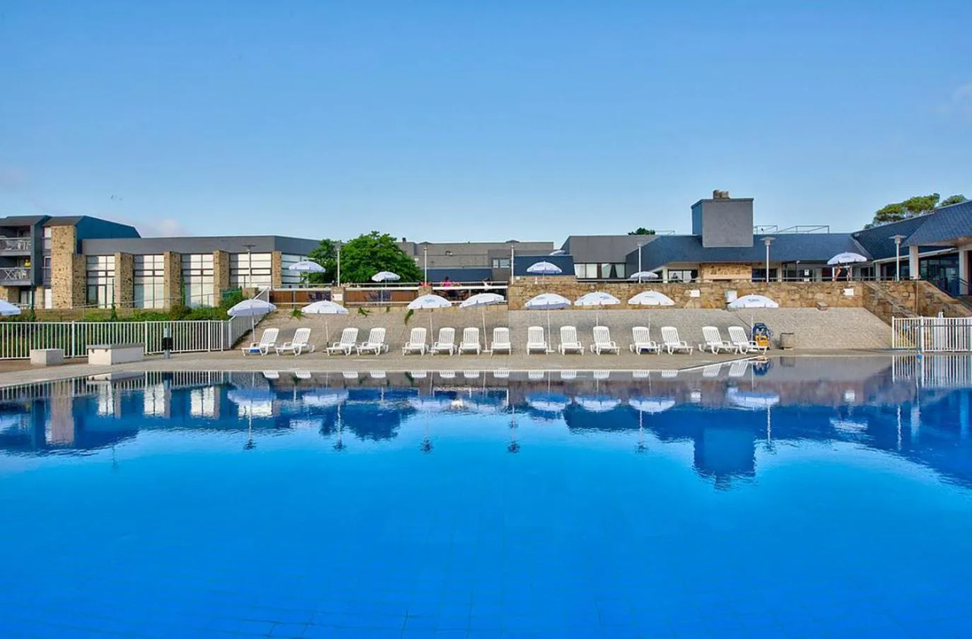 Une piscine bleue reflète un bâtiment avec des transats et des parasols.