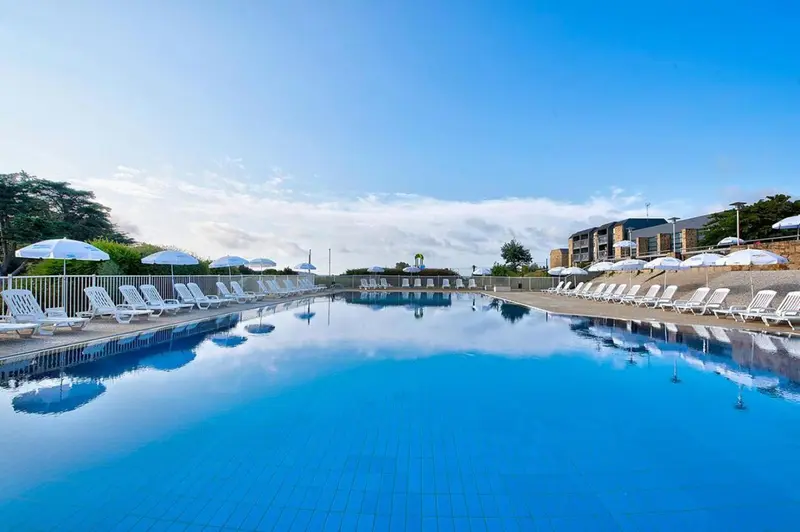 Une piscine rectangulaire bleu clair reflète le ciel et les parasols blancs.