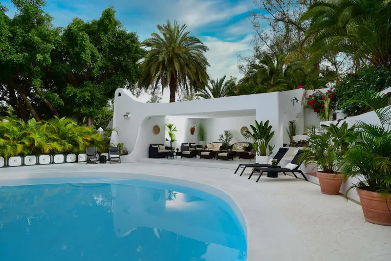Une piscine turquoise bordée de chaises longues et de végétation luxuriante devant un bâtiment blanc.