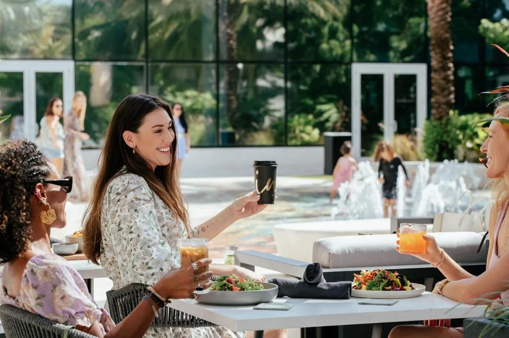 Des femmes discutent autour de tables en extérieur avec des boissons et de la nourriture.