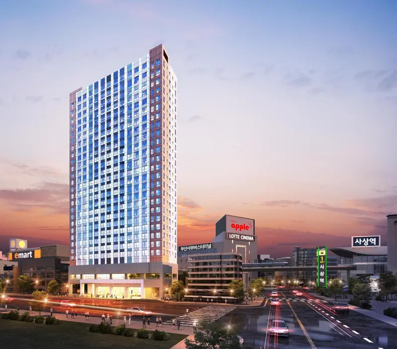 Immeuble moderne illuminé au crépuscule avec des commerces en rez-de-chaussée et une route passante.