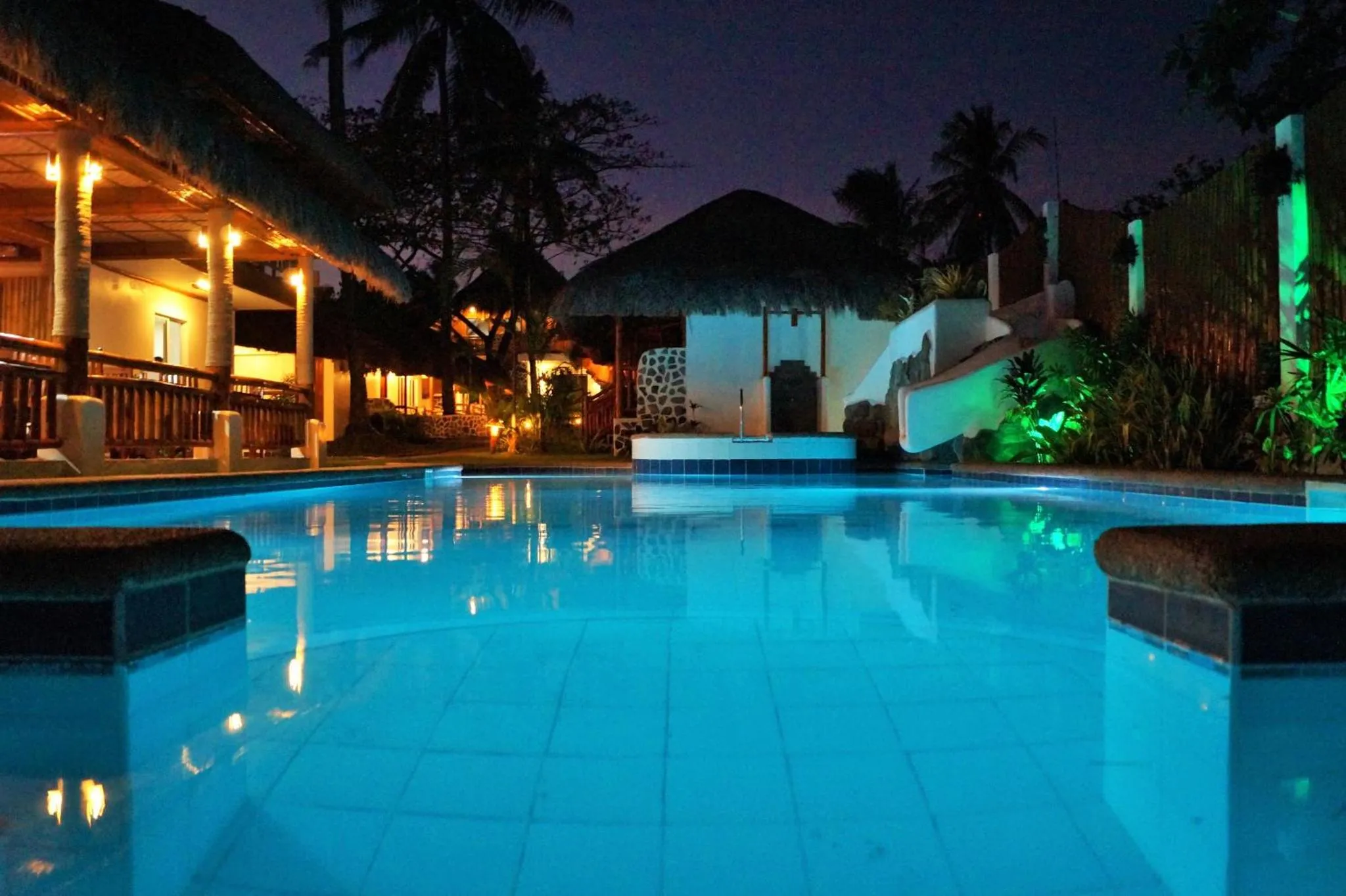 Piscine illuminée au crépuscule avec végétation tropicale.
