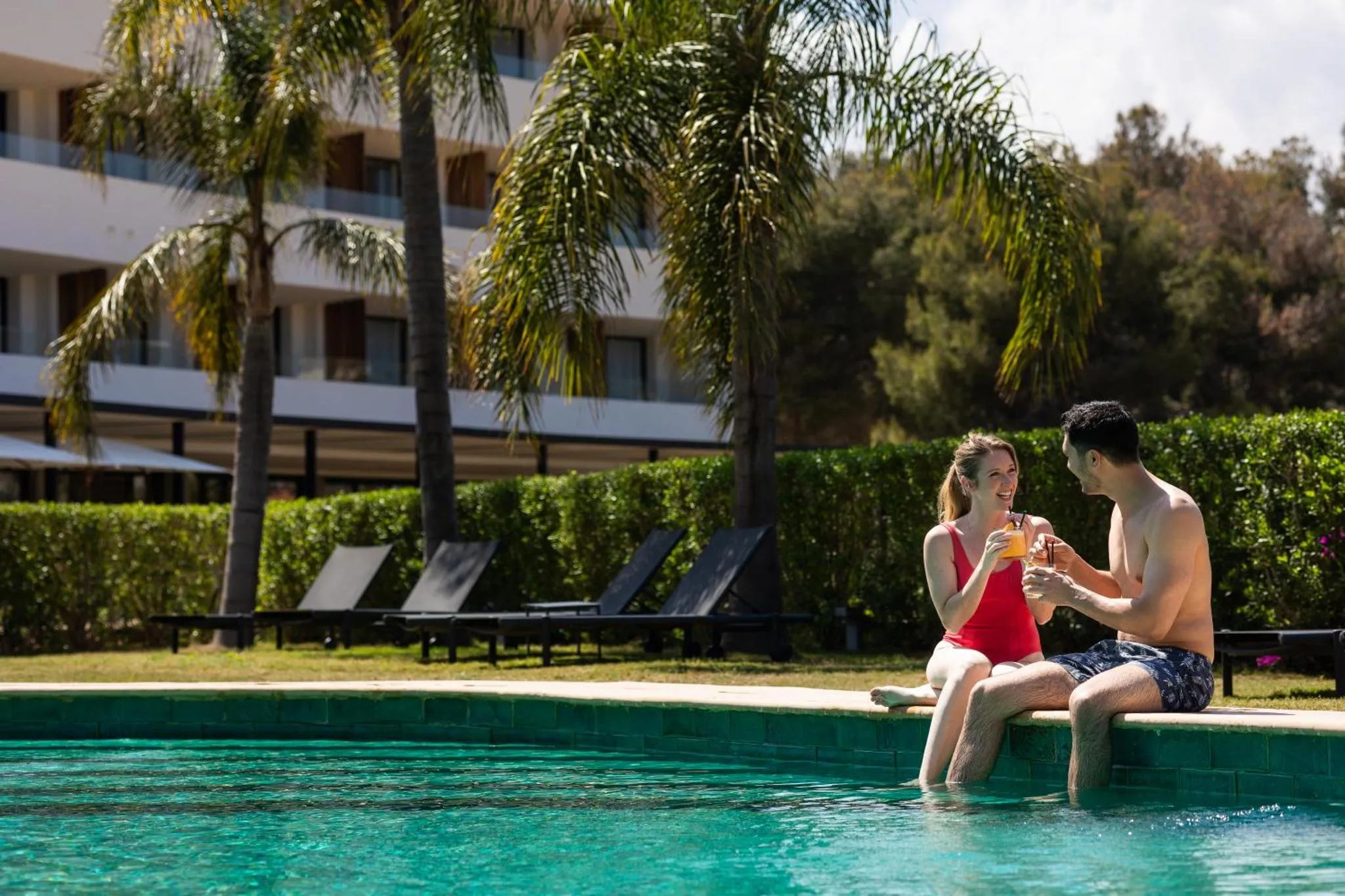Un couple souriant partage des boissons au bord d'une piscine ensoleillée.