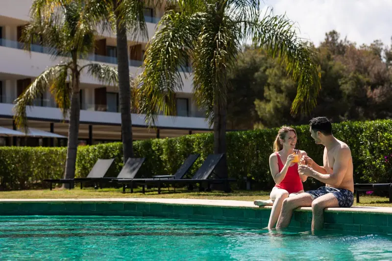 Un couple souriant partage des boissons au bord d'une piscine ensoleillée.