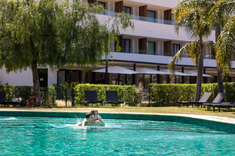 Une piscine bordée de transats et de végétation luxuriante devant un bâtiment moderne.