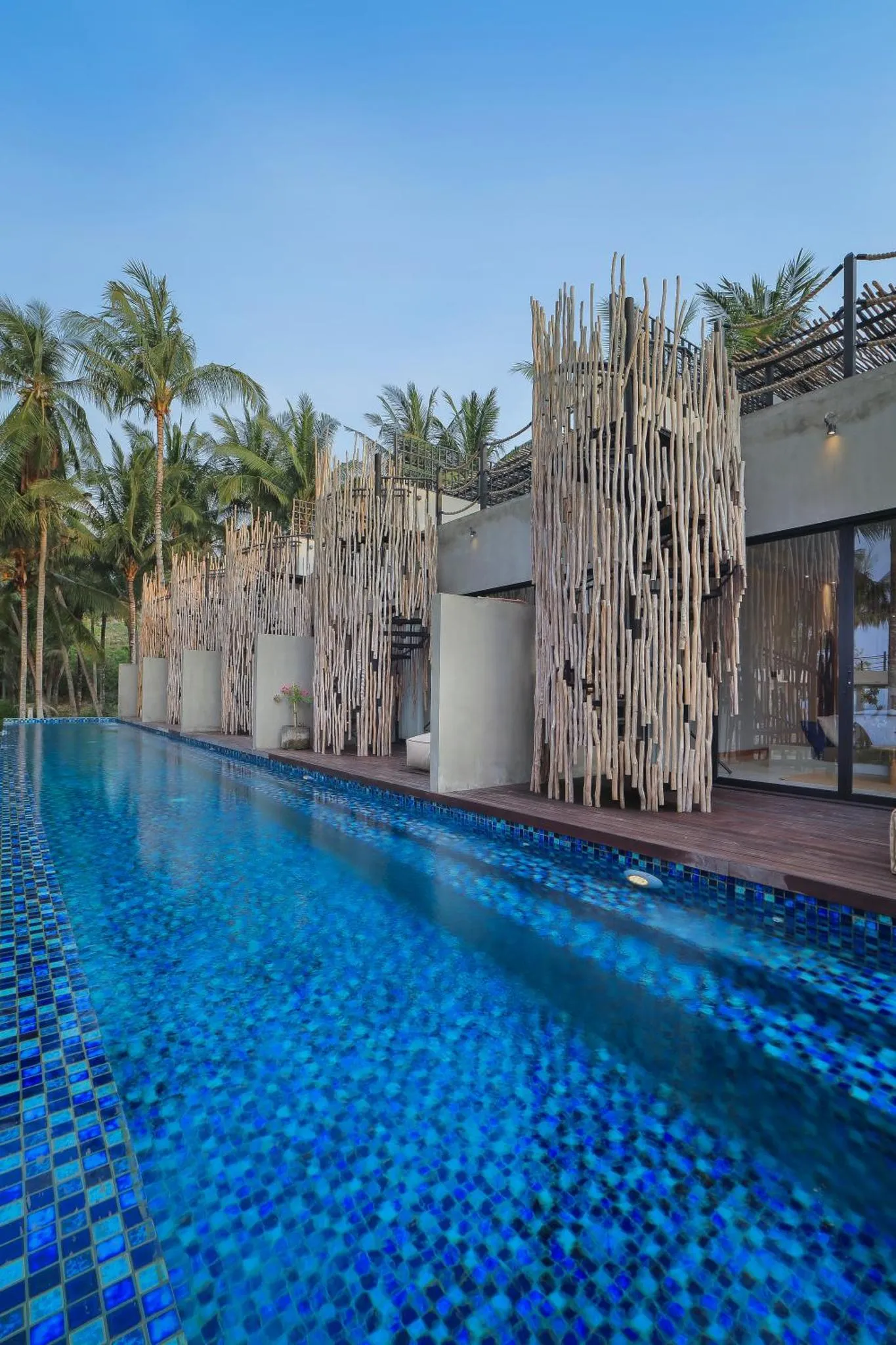 Une piscine rectangulaire aux carreaux bleus borde une terrasse en bois ombragée de palmiers.