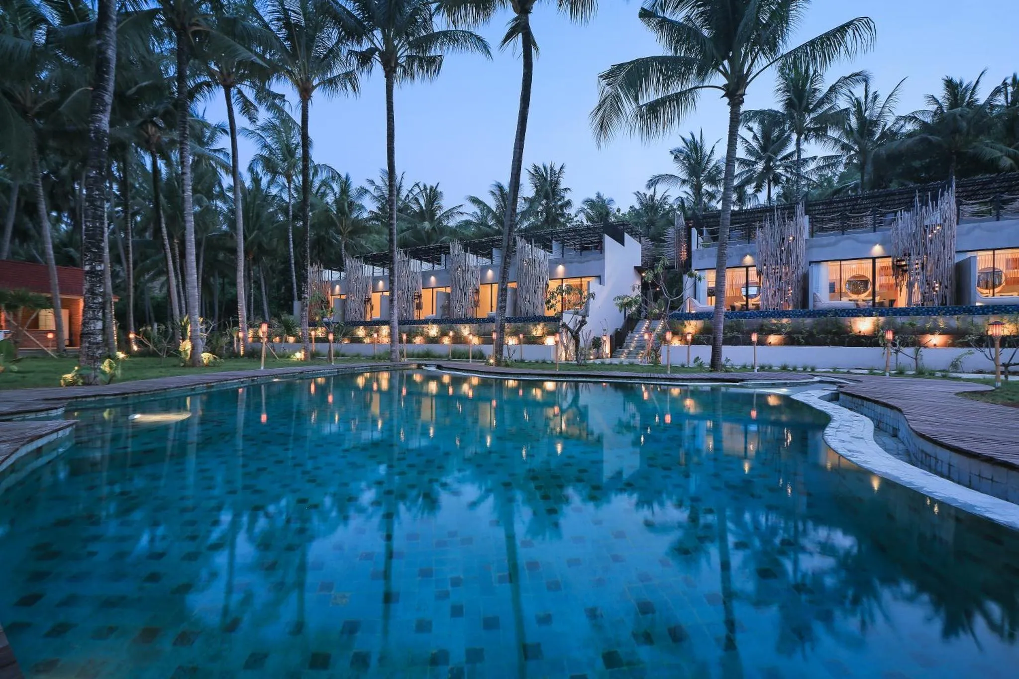 Une piscine turquoise reflète les lumières d'un bâtiment bordé de palmiers au crépuscule.
