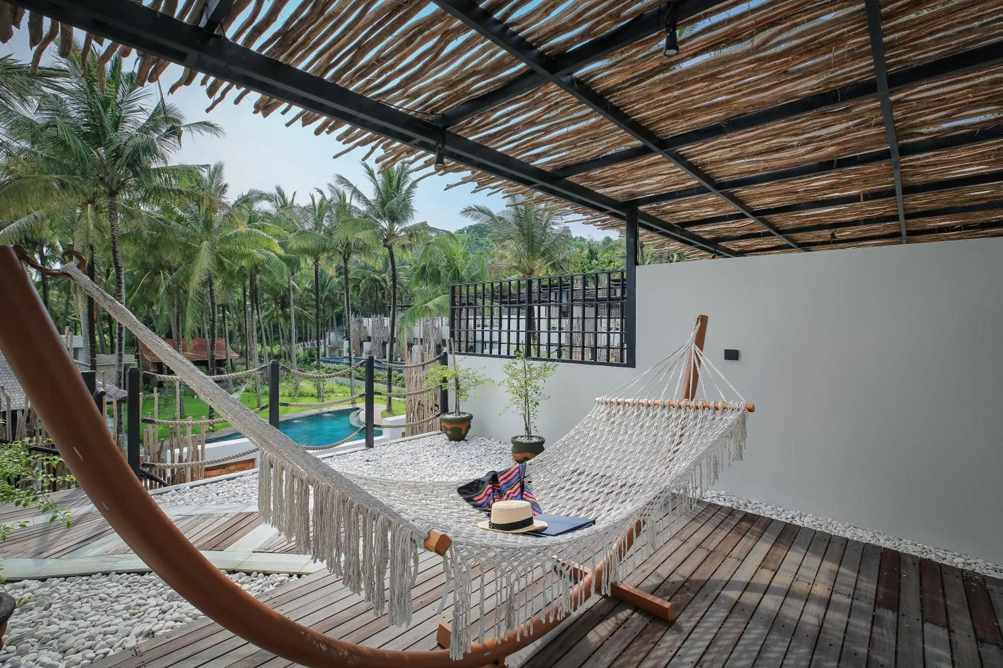 Un hamac est suspendu sur une terrasse en bois avec vue sur une piscine et une végétation tropicale.