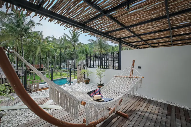 Un hamac est suspendu sur une terrasse en bois avec vue sur une piscine et une végétation tropicale.