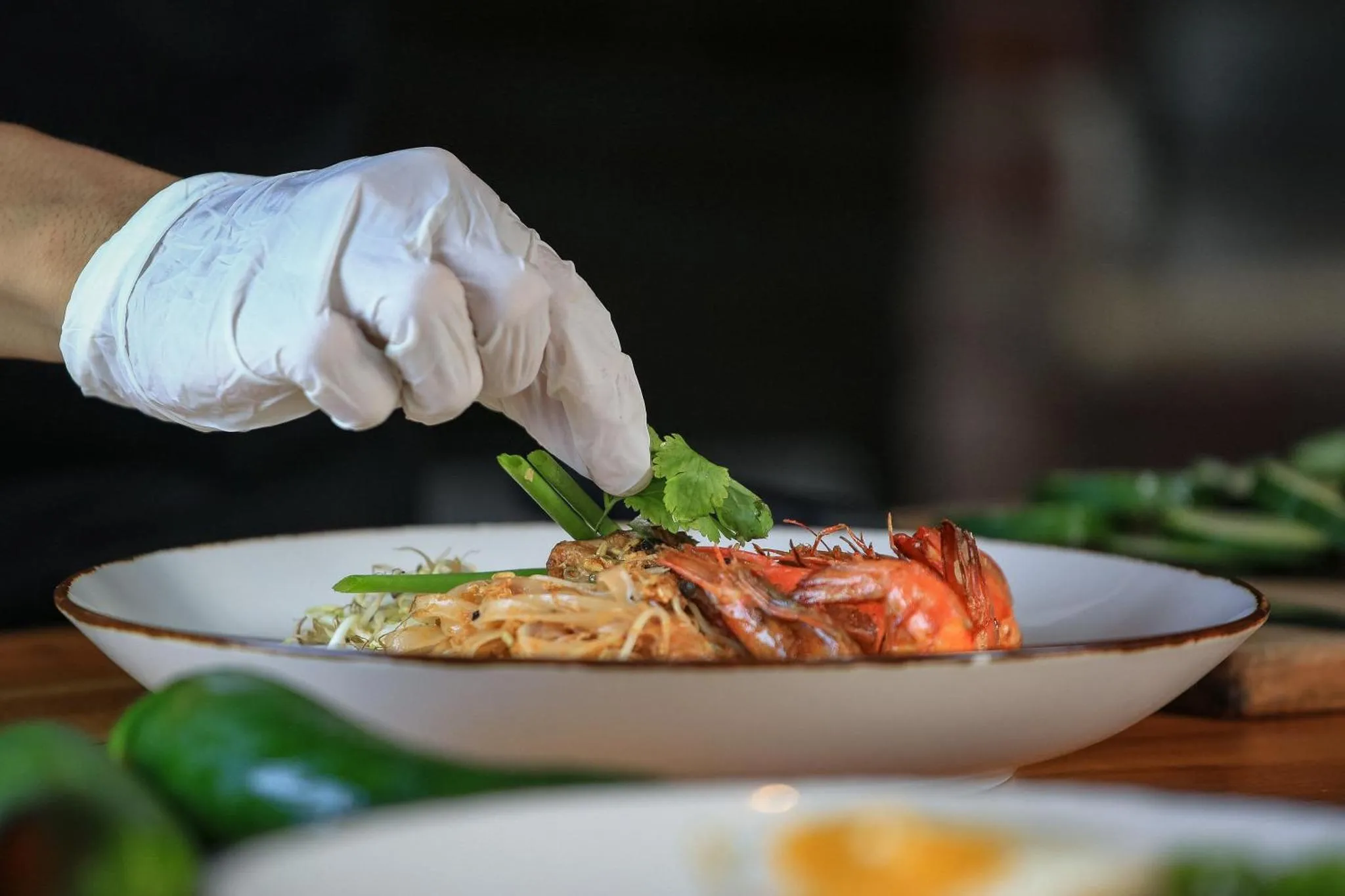 Une main gantée décore un plat de crevettes et de nouilles sur une assiette blanche.