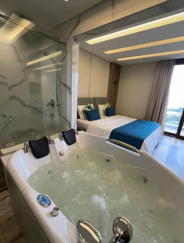 Une baignoire d'angle remplie d'eau se trouve dans une salle de bain moderne avec une douche à l'italienne et un lit en arrière-plan.