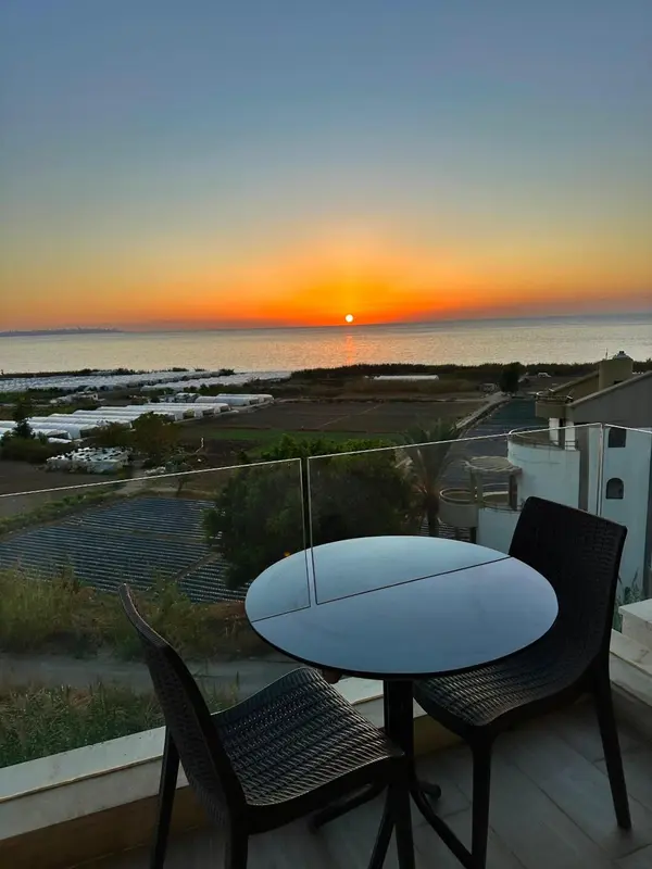 Une table ronde et deux chaises en osier sur une terrasse avec vue sur la mer au coucher du soleil.