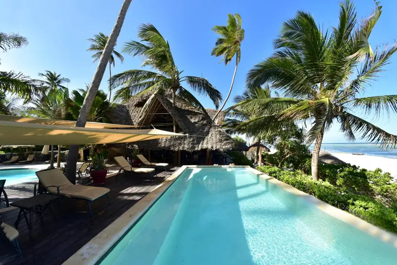 Transats et piscine turquoise bordent une plage de sable blanc sous les cocotiers.