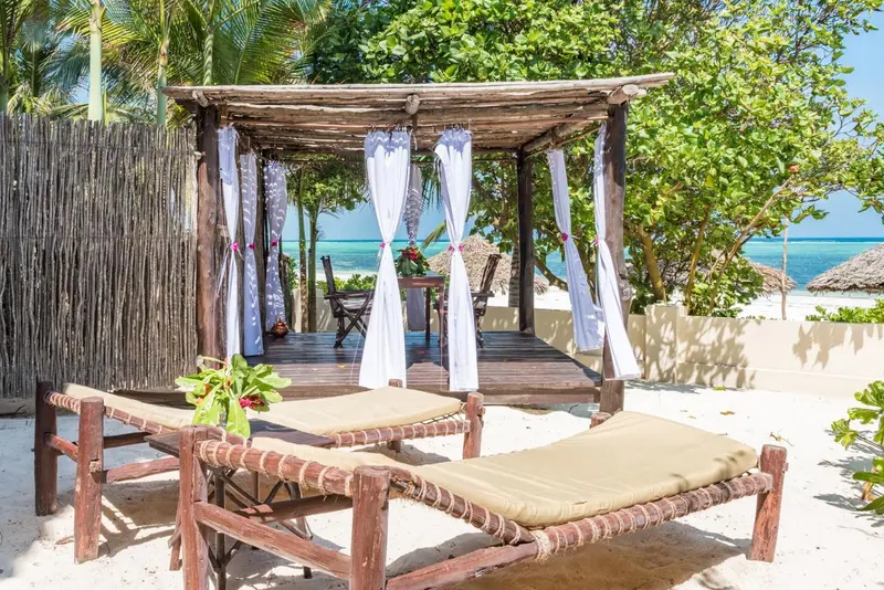 Deux chaises longues en bois sur une plage de sable blanc, sous un abri ajouré décoré de voilages.