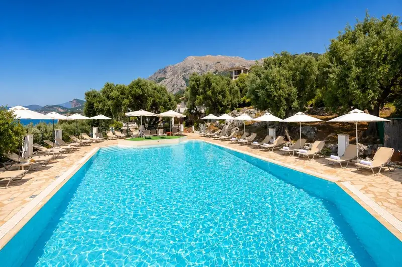 Une piscine rectangulaire bordée de transats sous des parasols, avec une vue sur la mer et des montagnes.