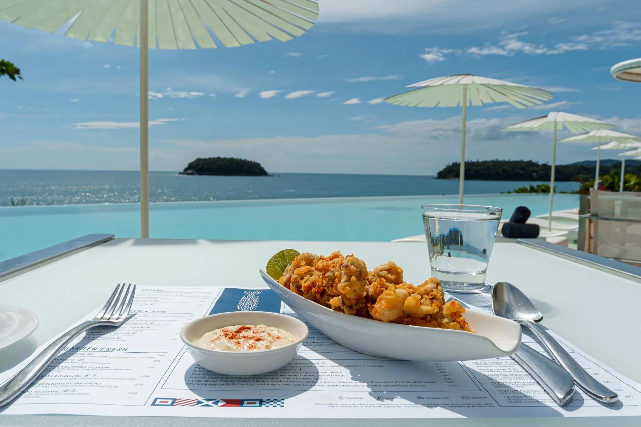 Une table est dressée avec des plats de fruits de mer et des boissons, face à une piscine à débordement et une vue sur la mer.