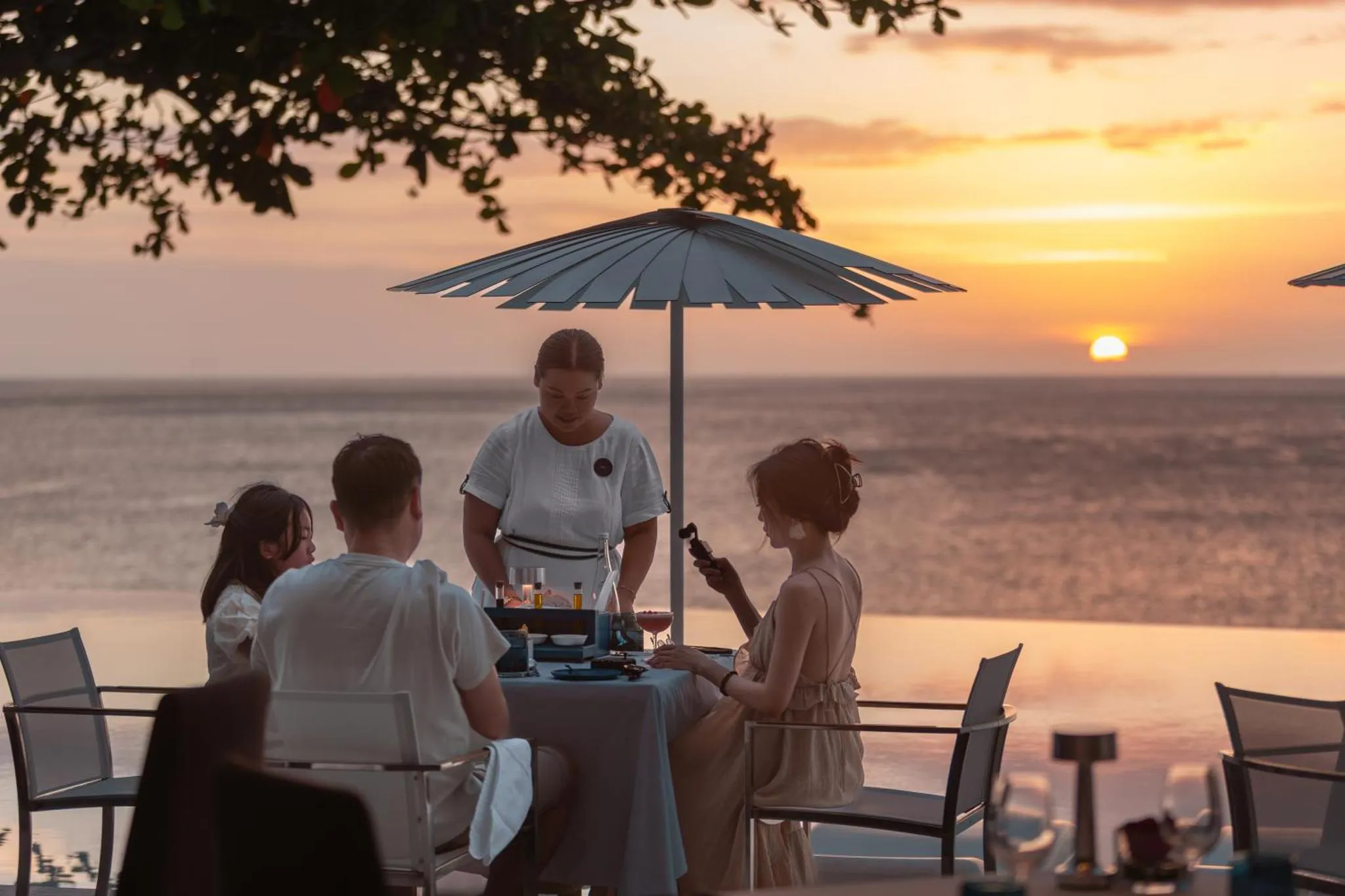 Des convives sont servis à table au bord d'une piscine face à la mer au coucher du soleil.