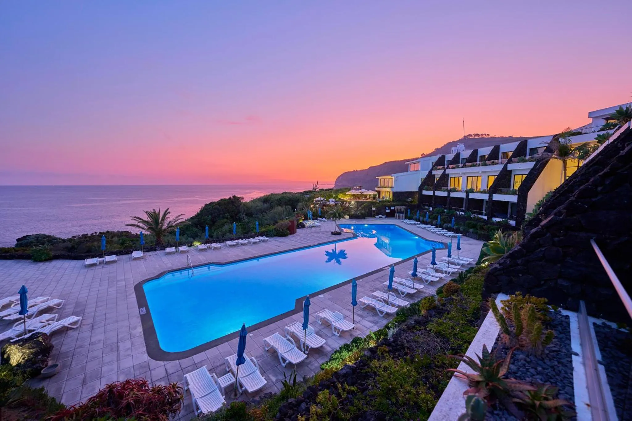 Une piscine rectangulaire avec des transats au bord, devant un bâtiment et face à la mer au coucher du soleil.