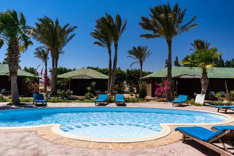 Une piscine turquoise entourée de chaises longues et de bâtiments aux toits de chaume sous des palmiers.