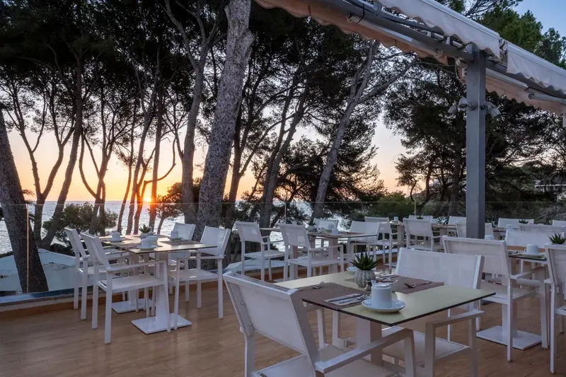 Tables et chaises blanches dressées sur une terrasse en bois face à la mer au coucher du soleil.