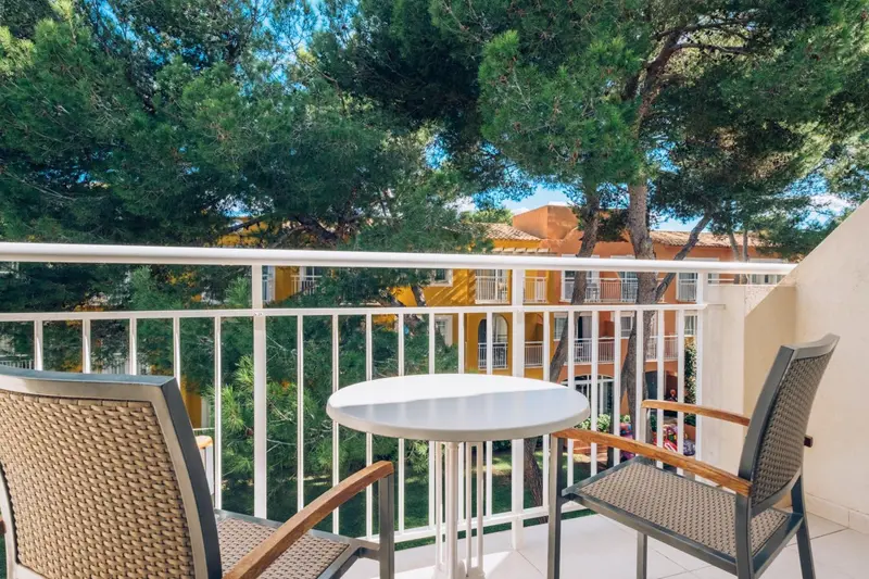 Une terrasse avec une table ronde et deux chaises en osier offre une vue sur un bâtiment jaune et des arbres verts.