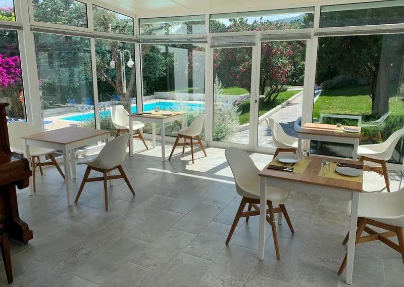 Une salle lumineuse avec des tables et des chaises blanches, donnant sur une piscine et un jardin.