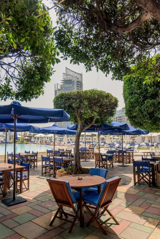 Tables et chaises bleues sous des parasols dans un espace extérieur arboré.