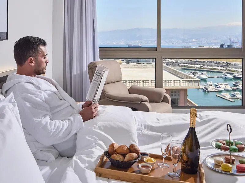 Un homme lit un journal assis sur un lit dans une chambre avec vue sur un port.
