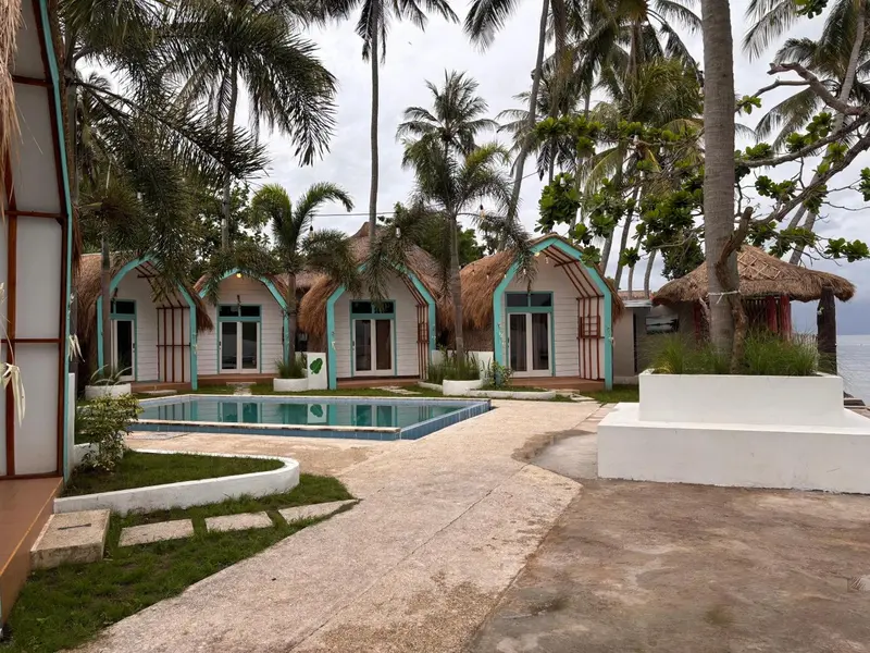 Petites maisons de vacances turquoise et blanches avec toits de chaume bordent une piscine rectangulaire.