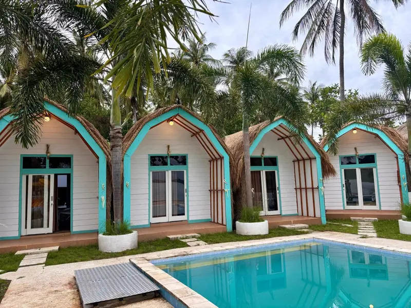 Petites maisons de vacances turquoise et blanches avec toits de chaume bordent une piscine rectangulaire.